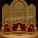 "Luxurious Mughal wedding stage with golden metal backdrop frames, intricate lattice patterns, red velvet sofa and chairs, and royal event décor setup."