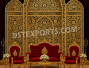 "Luxurious Mughal wedding stage with golden metal backdrop frames, intricate lattice patterns, red velvet sofa and chairs, and royal event décor setup."